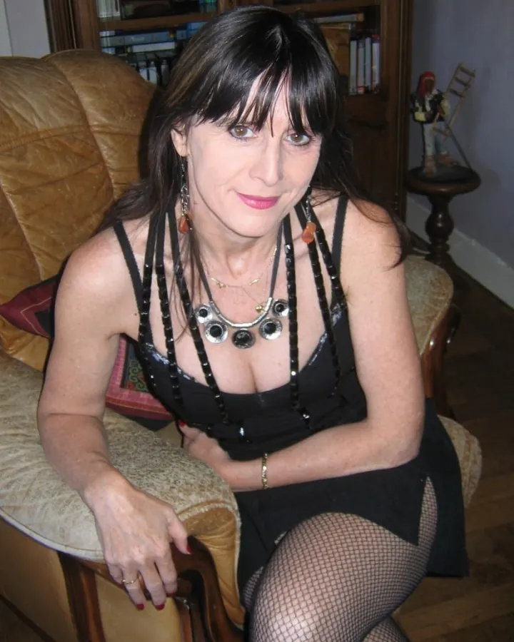 Femme aux cheveux bruns-noirs, assise sur un canapé en cuir, portant une robe noire avec des chaînes et des collants à mailles.