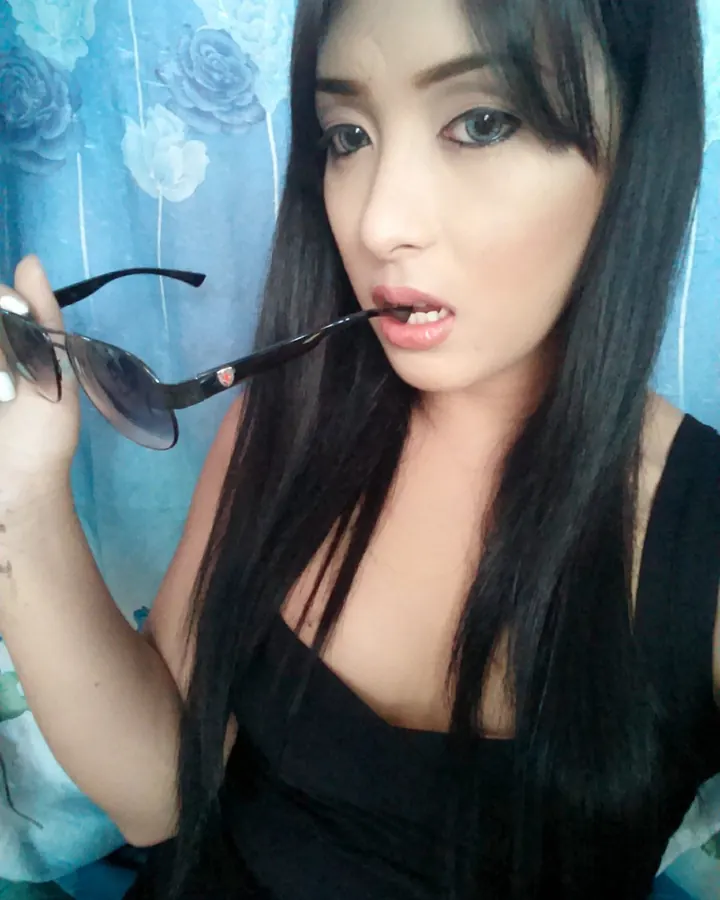 Jeune femme asiatique aux cheveux bruns-noirs, vêtue d'un haut noir, tenant des lunettes de soleil dans sa bouche.