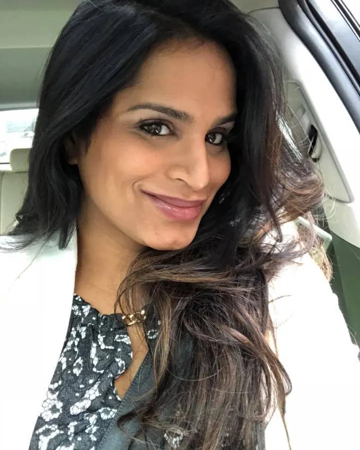 Femme aux cheveux foncés, souriante, assise dans une voiture, vêtue d'une veste blanche et d'un haut imprimé.