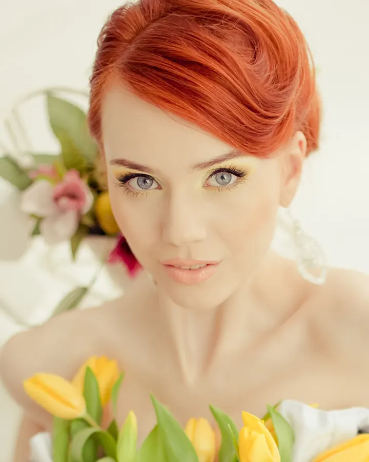 Femme aux cheveux roux, yeux bleus, tenant des tulipes jaunes, maquillage lumineux, fond blanc.