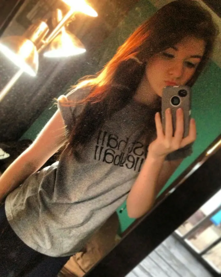 Femme aux cheveux longs foncés, prenant un selfie dans un miroir, portant un t-shirt gris et regardant son téléphone.