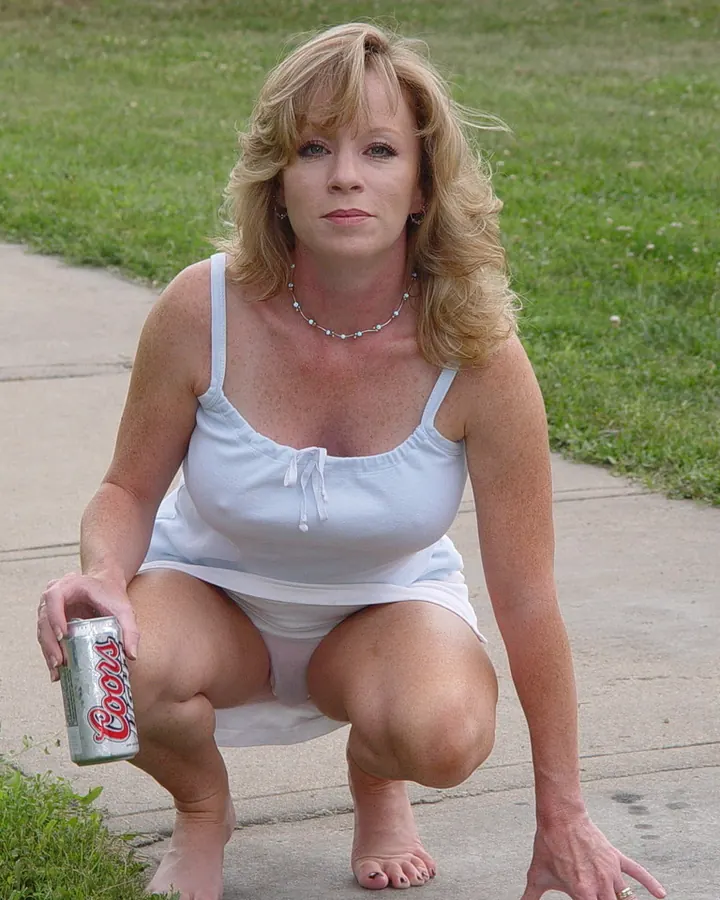 Femme blonde en robe blanche, accroupie sur un trottoir, tenant une canette de bière, regardant l'objectif.