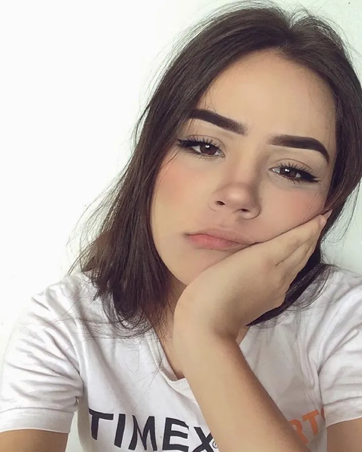 Jeune femme aux cheveux bruns portant un t-shirt blanc avec l'inscription 'TIMEX', posant avec la main sur le visage contre un fond blanc.
