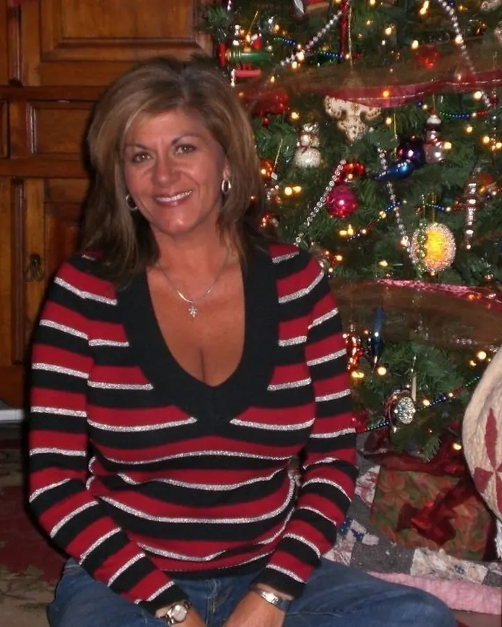 Femme souriante aux cheveux bruns, portant un pull rayé rouge, noir et blanc, assise devant un sapin de Noël décoré.