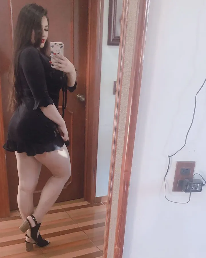 Femme blonde aux cheveux châtains, portant un haut rose et une jupe noire, prenant un selfie devant un miroir dans un couloir.