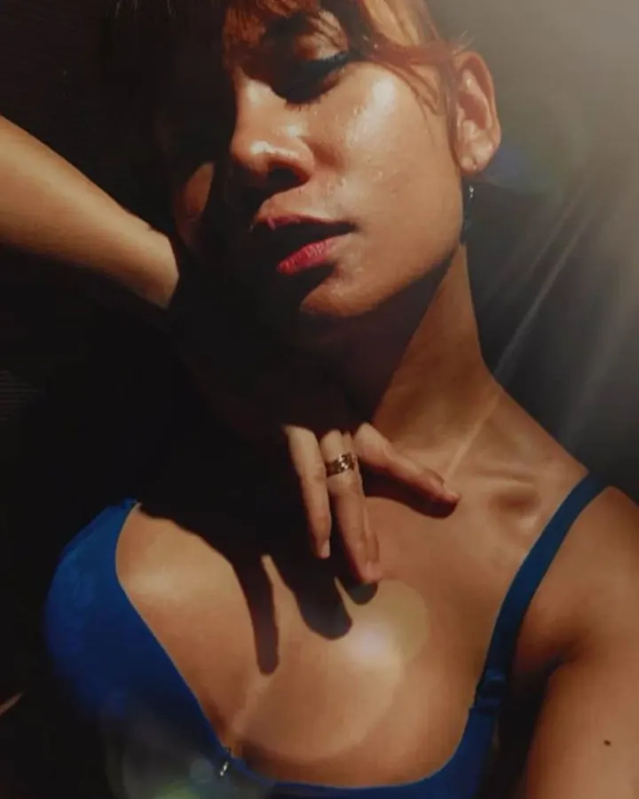 Plan rapproché d'une femme aux cheveux châtains et yeux marrons, portant un soutien-gorge bleu, la main sur le cou, sous un éclairage dramatique.
