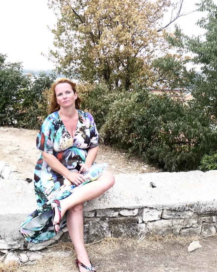 Femme aux cheveux roux assise sur un mur en pierre, vêtue d'une robe colorée à motifs, regardant l'objectif.