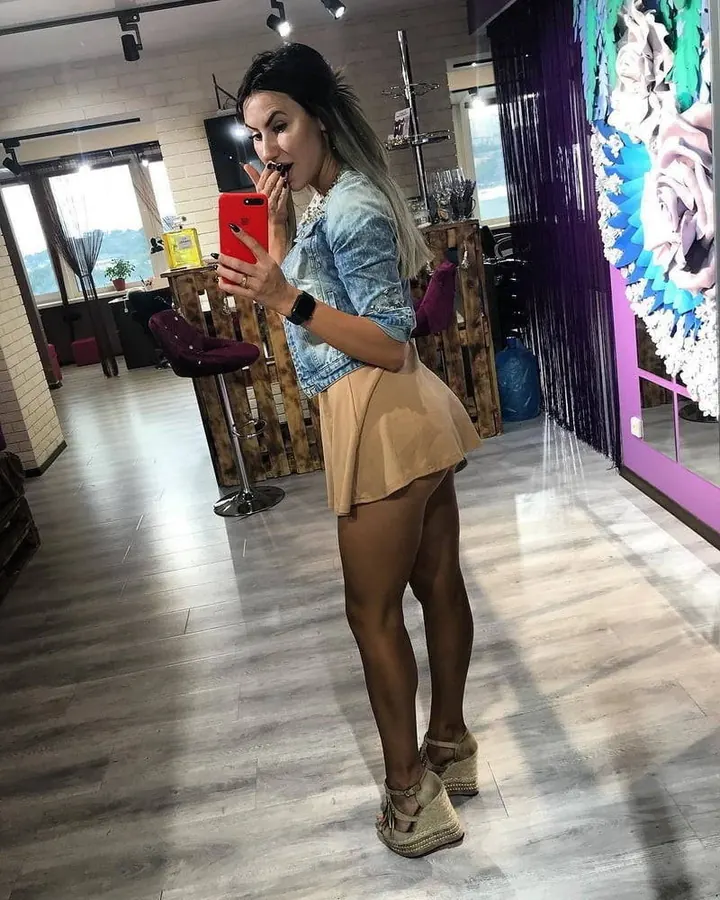 Femme arabe aux cheveux bruns-noirs, portant une tenue noire avec des gants, posant dans un intérieur aux murs violets.