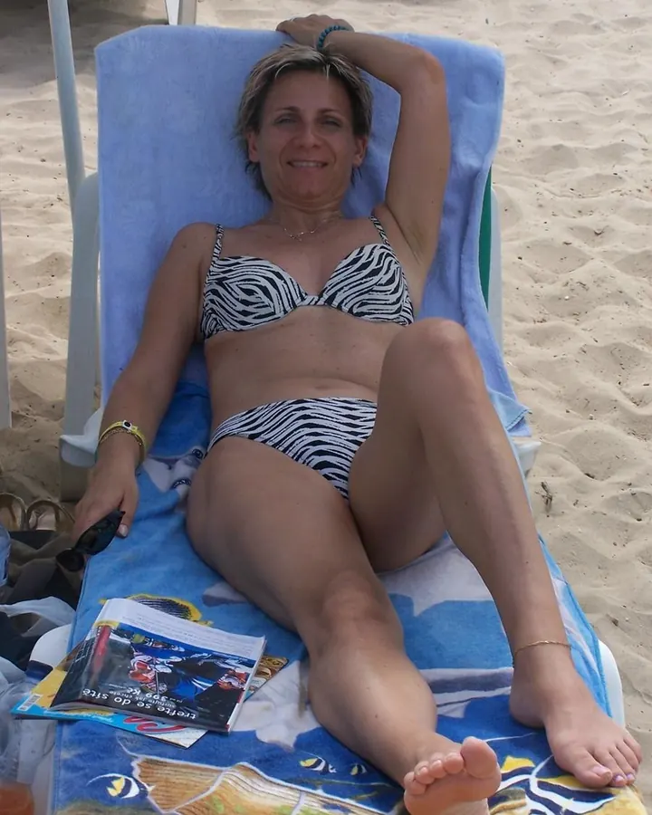 Femme aux cheveux blonds, en robe bleue, assise sur un canapé, souriante, main sur le menton.