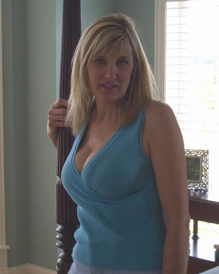 Femme aux cheveux blonds portant un haut bleu, debout près d'une rampe en bois dans une pièce, regardant l'appareil.