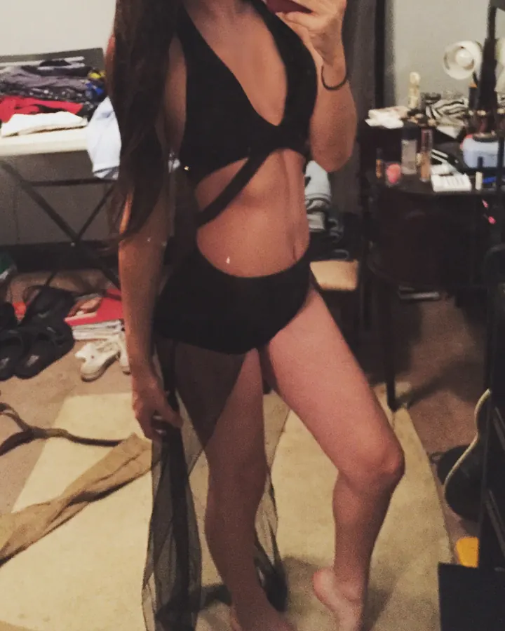 Femme aux cheveux bruns portant un body noir à découpes, tenant un tissu noir dans une chambre en désordre.