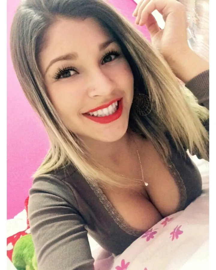 Femme aux cheveux blonds portant un haut rayé bleu et blanc, un jean déboutonné, debout dans un jardin, regardant l'appareil.