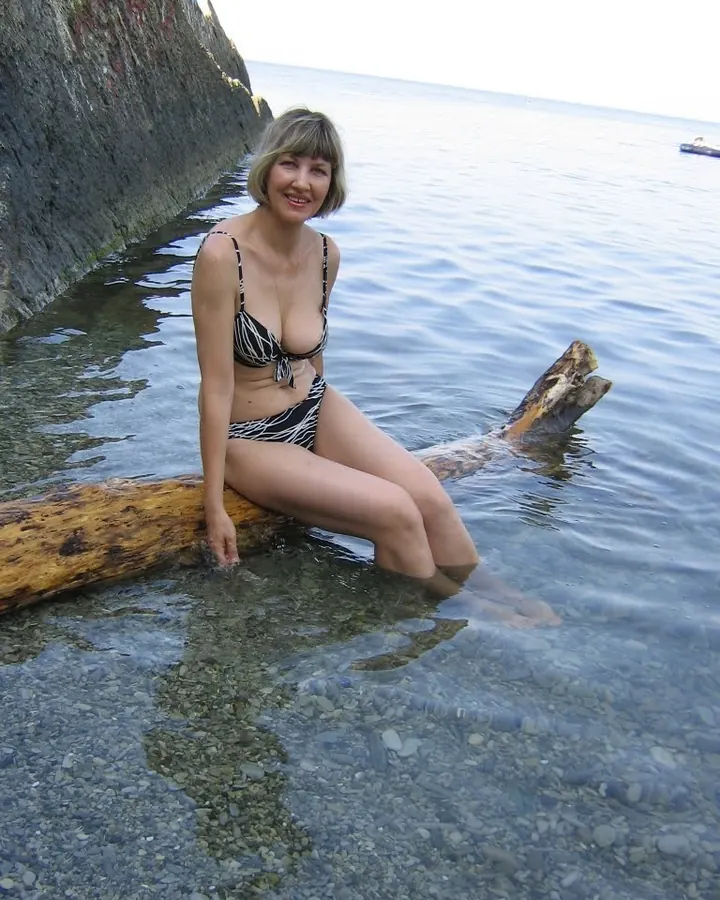 Femme aux cheveux blonds portant un bikini noir et blanc, assise sur un tronc d'arbre dans l'eau claire, souriant vers l'appareil.
