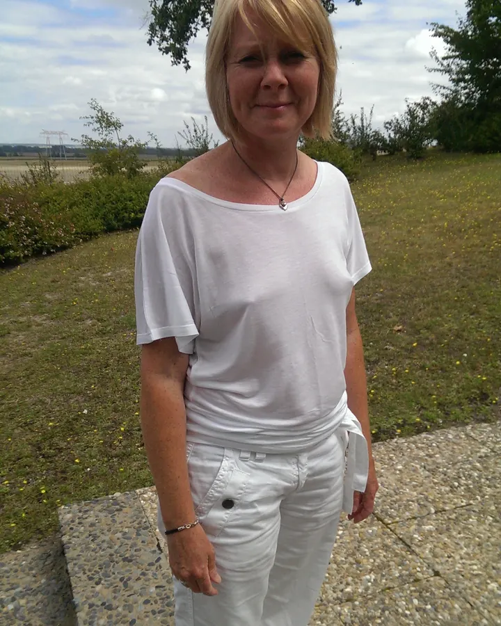Femme blonde en tenue blanche, debout en extérieur, souriant, avec un ciel nuageux et des arbres en arrière-plan