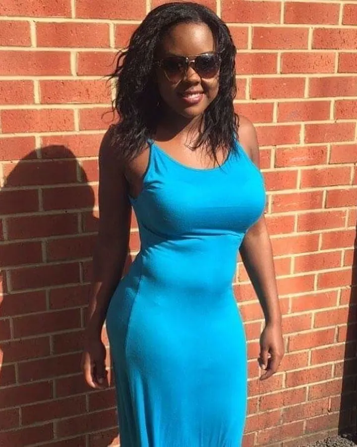 Femme africaine aux cheveux noirs en robe bleue ajustée, souriant, devant un mur de briques rouges