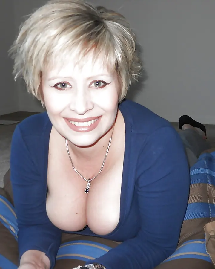 Femme aux cheveux platine courts, souriante, vêtue d'un haut bleu décolleté, assise sur un canapé rayé dans une pièce aux murs gris.