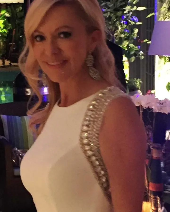 Femme blonde aux cheveux mi-longs, souriante, vêtue d'une robe blanche ornée de strass, debout dans un décor verdoyant et lumineux.