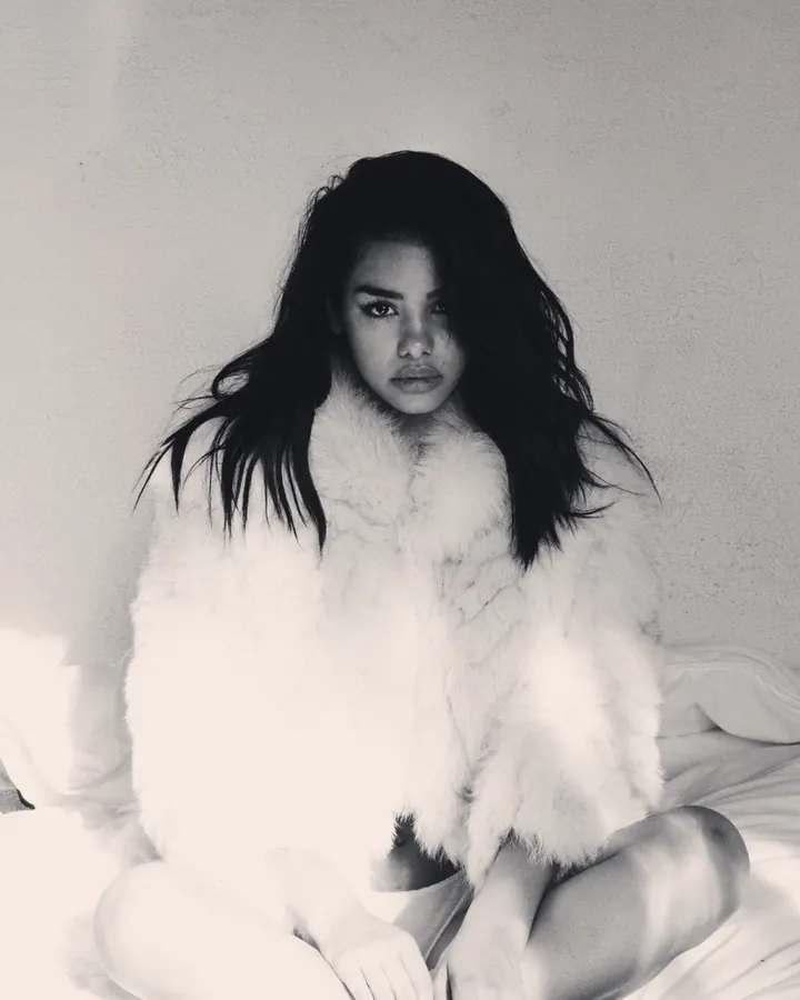 Femme aux cheveux noirs longs, assise sur un lit, portant une fourrure blanche, dans une photo en noir et blanc.