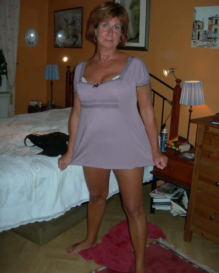 Femme aux cheveux roux mi-longs, souriante, vêtue d'un haut rose, assise sur un fauteuil dans une chambre.