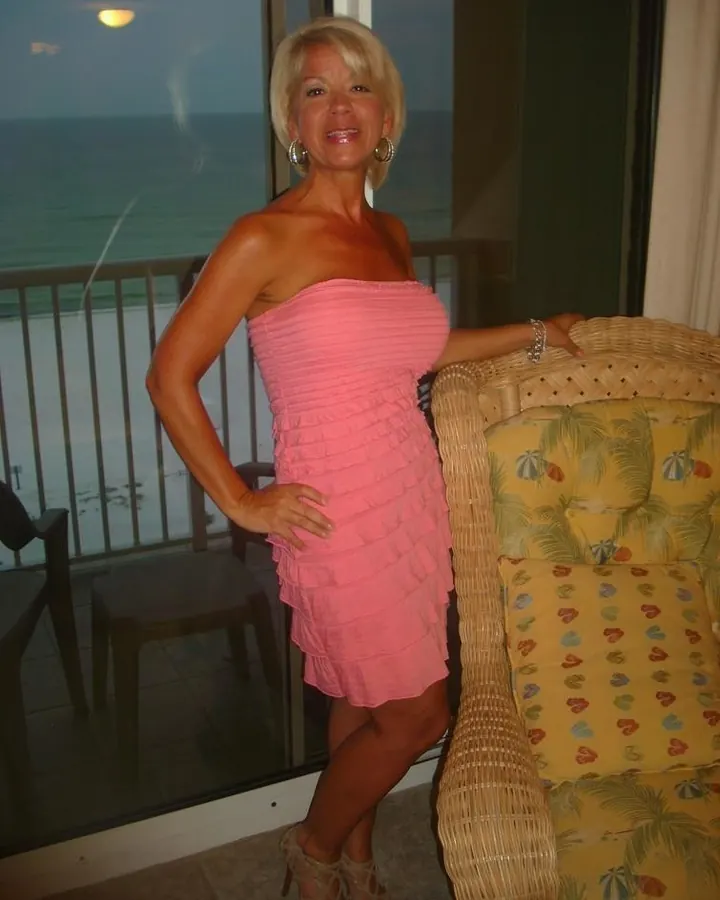 Femme souriante en robe rose sans bretelles, debout sur un balcon avec vue sur la mer, portant des sandales à talons et des boucles d'oreilles dorées.