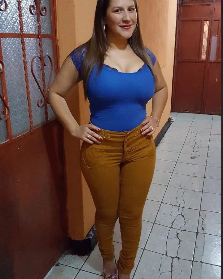 Femme aux cheveux bruns portant un haut bleu et un pantalon marron, souriante, les mains sur les hanches.