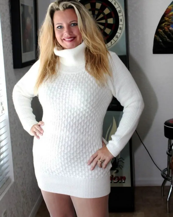 Femme blonde aux cheveux platine, souriante, portant un pull blanc, les mains sur les hanches devant un jeu de fléchettes.