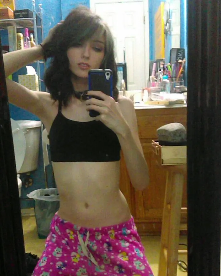 Femme en haut noir et pantalon rose à motifs, prenant un selfie dans une salle de bain bleue.