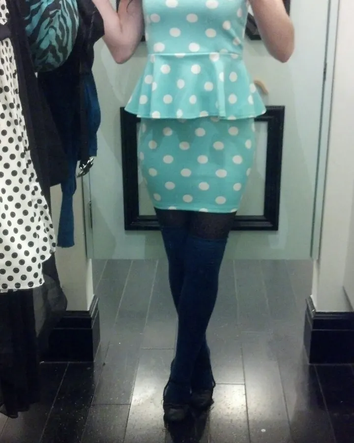Femme en robe à pois turquoise, collants noirs et chaussures noires, prenant un selfie devant un miroir.