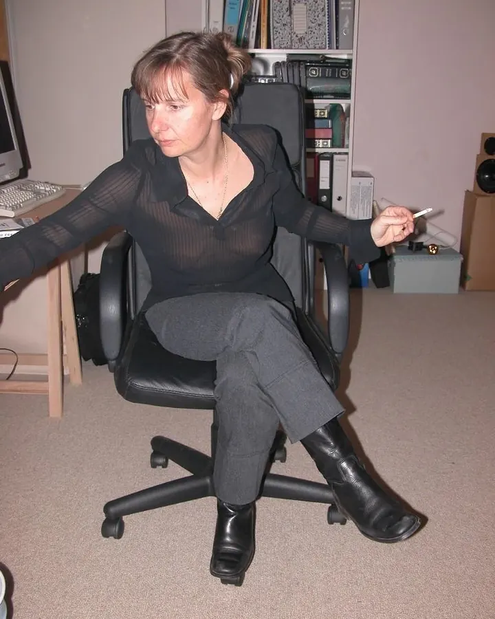Femme en chemise noire transparente, assise dans un fauteuil de bureau, tenant une cigarette.