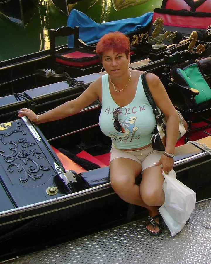 Femme aux cheveux roux en haut de couleur claire, assise sur un bateau noir orné de dorures.