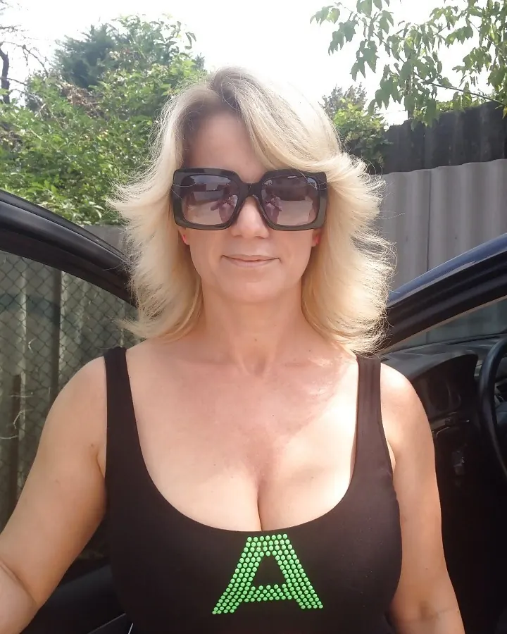 Femme blonde portant des lunettes de soleil et un haut noir avec des lettres colorées, debout près d’une voiture.
