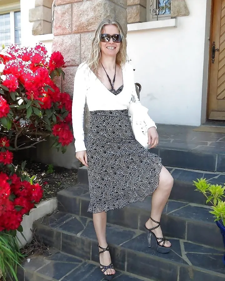 Femme blonde, portant une robe blanche et une jupe à motifs, assise sur des marches extérieures, souriant.