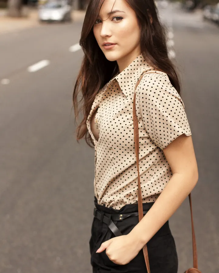 Femme aux cheveux bruns en chemise à pois beige et jupe noire, marchant dans une rue urbaine avec un sac en cuir marron.