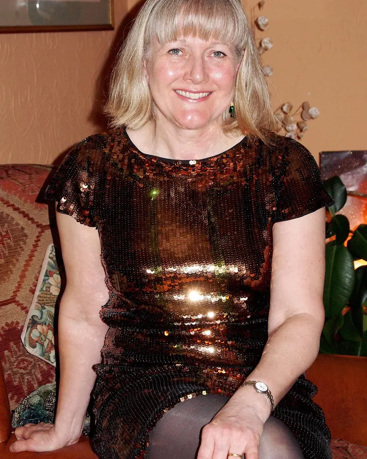 Femme aux cheveux blonds platine, portant une robe noire en dentelle, assise sur un canapé doré, souriant à la caméra.