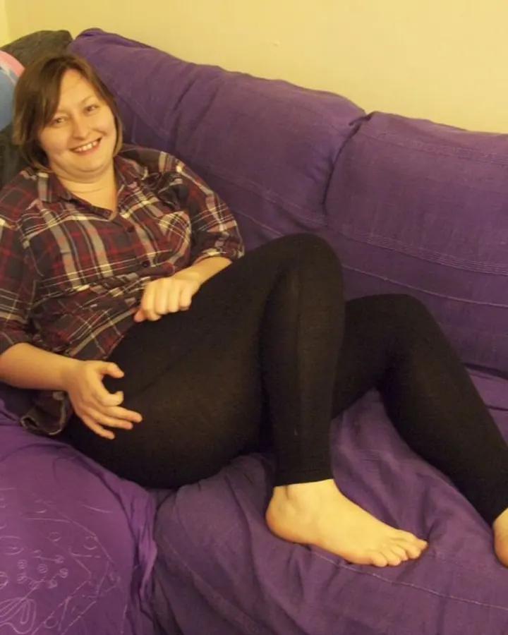 Femme aux cheveux châtains, portant une chemise à carreaux et un legging noir, assise sur un canapé violet, souriant.