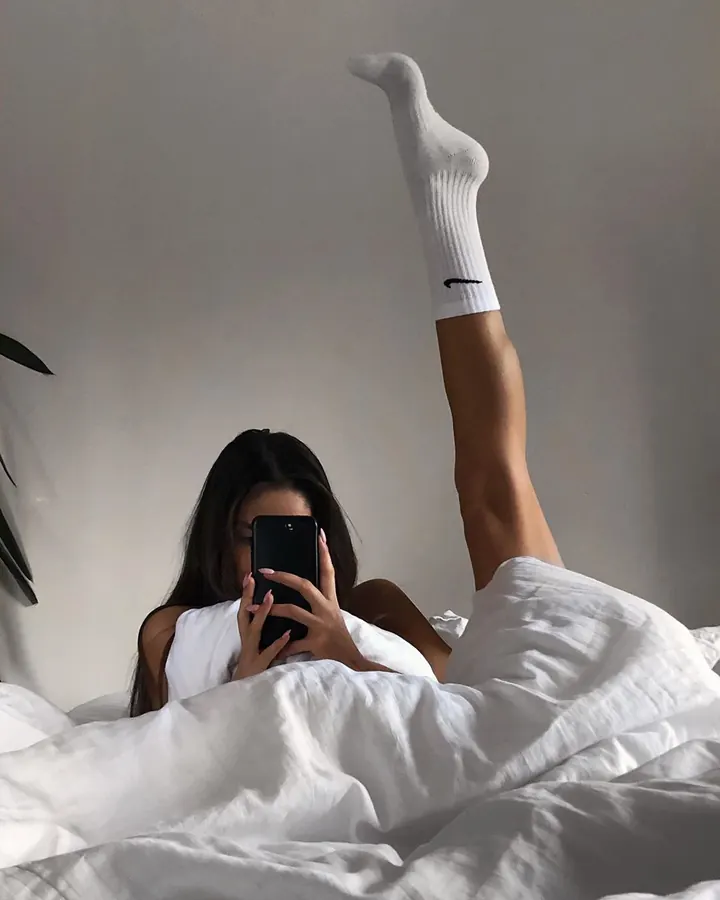 Jeune femme aux cheveux bruns noirs, portant un haut blanc et un short noir, allongée sur un lit blanc, enchaussée de chaussettes blanches.