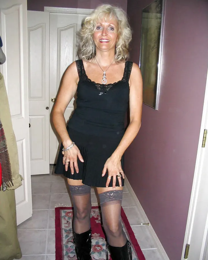 Femme aux cheveux blonds platine portant une robe noire à dentelle, des collants à motifs et des bottes noires, posant debout dans un couloir.