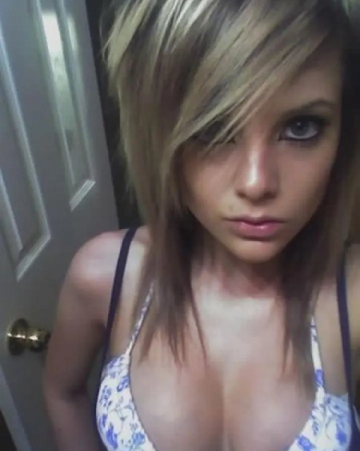 Femme aux cheveux blonds portant un haut blanc et un short noir, prenant un selfie dans une salle de bain.
