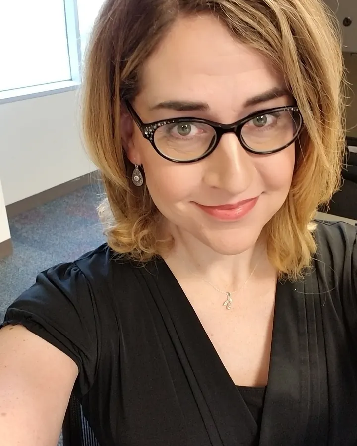 Femme aux cheveux bruns portant une robe noire, des lunettes et un collier, prenant un selfie en bureau.