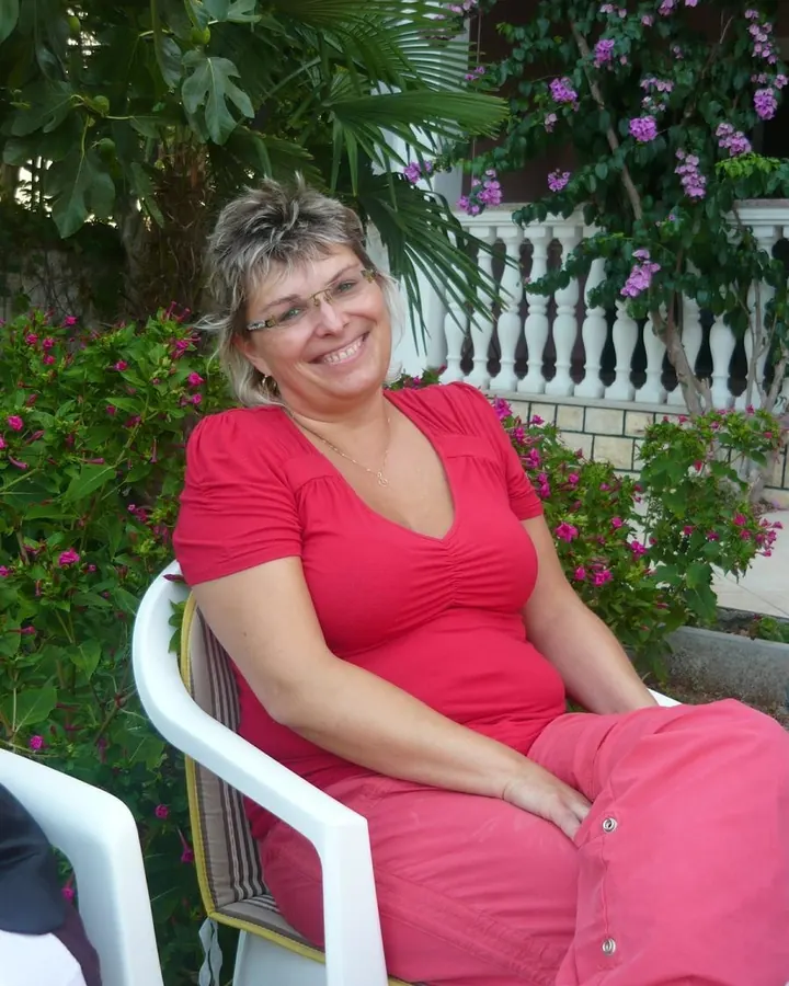 Femme aux cheveux blonds courts, portant des lunettes et une robe rouge, assise sur une chaise blanche dans un jardin fleuri.