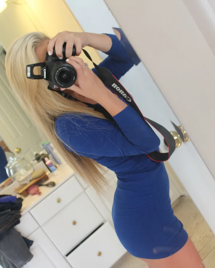 Femme aux cheveux blonds longs, portant une robe bleue ajustée, tenant un appareil photo Canon devant un miroir dans une salle de bain blanche.