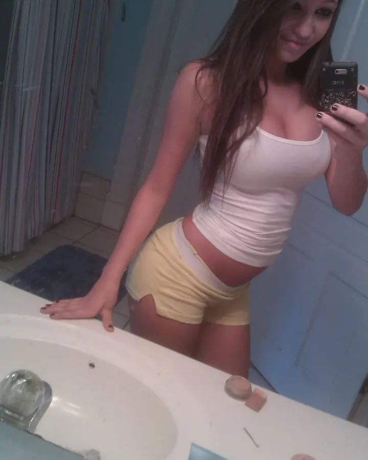 Jeune femme aux cheveux bruns, vêtue d'un haut blanc et d'un short jaune, prenant un selfie devant un miroir dans une salle de bain.