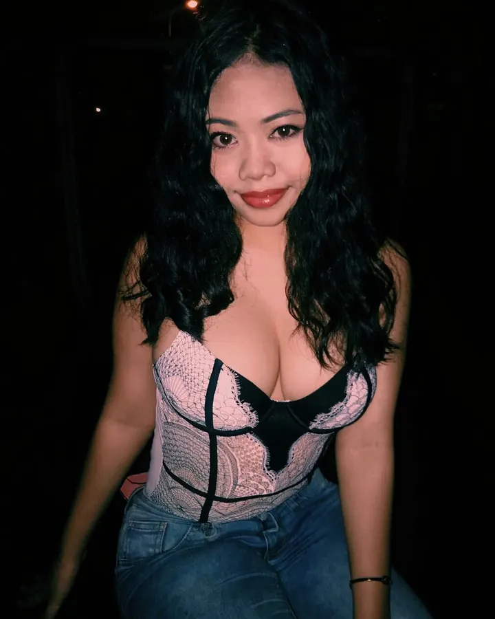 Jeune femme asiatique aux cheveux noirs, vêtue d'un haut de lingerie rose et noir, assise dans une ambiance sombre.