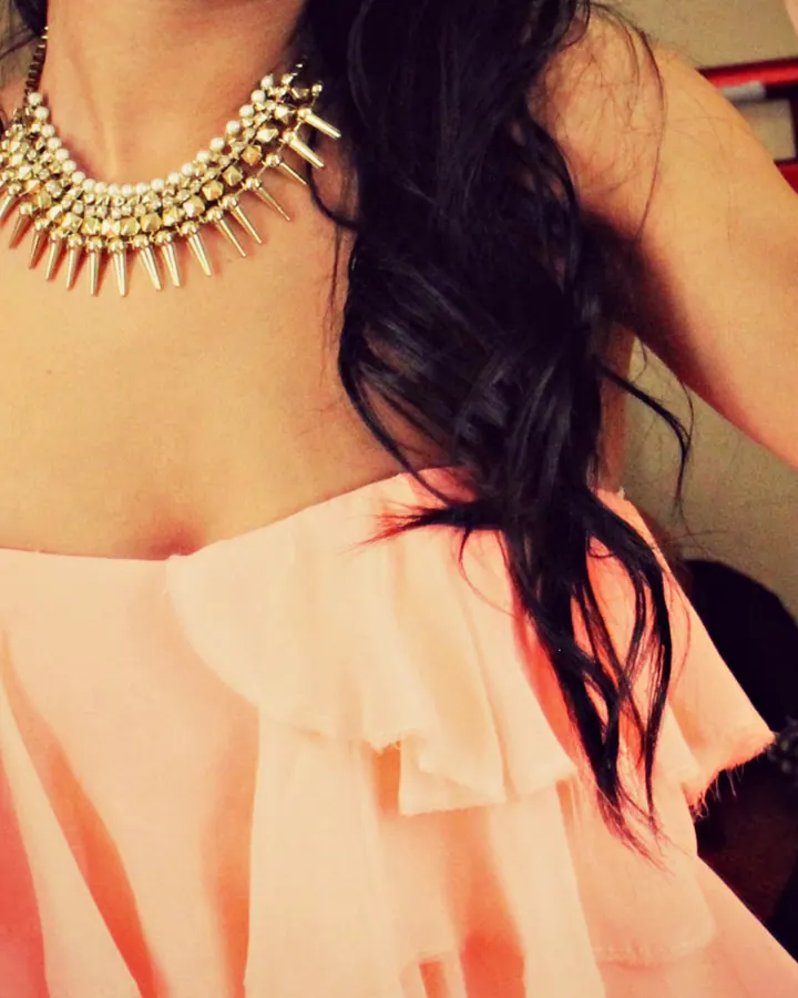 Femme en haut orange décolleté, portant un collier doré pointu, cheveux noirs ondulés.