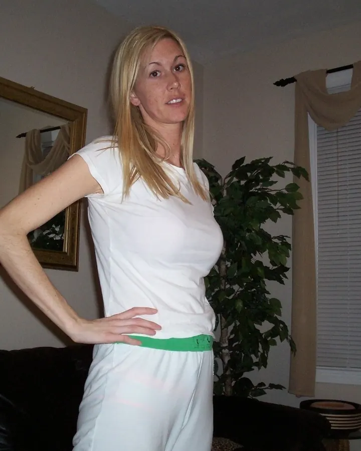 Femme blonde en t-shirt blanc et jupe blanche, main sur la hanche, dans un salon intérieur.