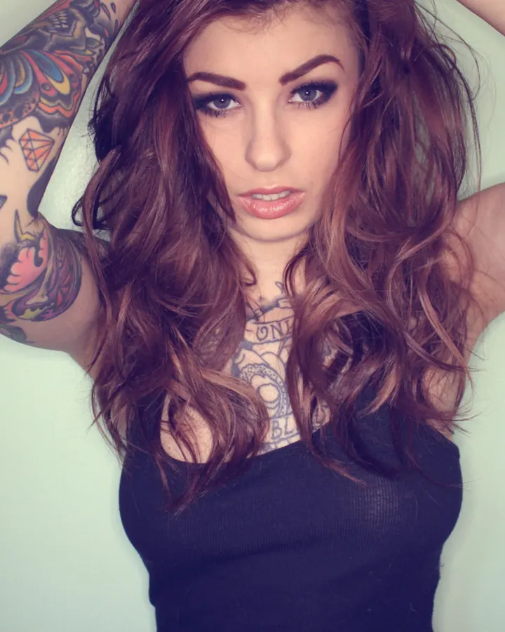 Femme aux cheveux châtains et yeux bleus, tatouée, portant un haut noir, les bras levés derrière la tête.
