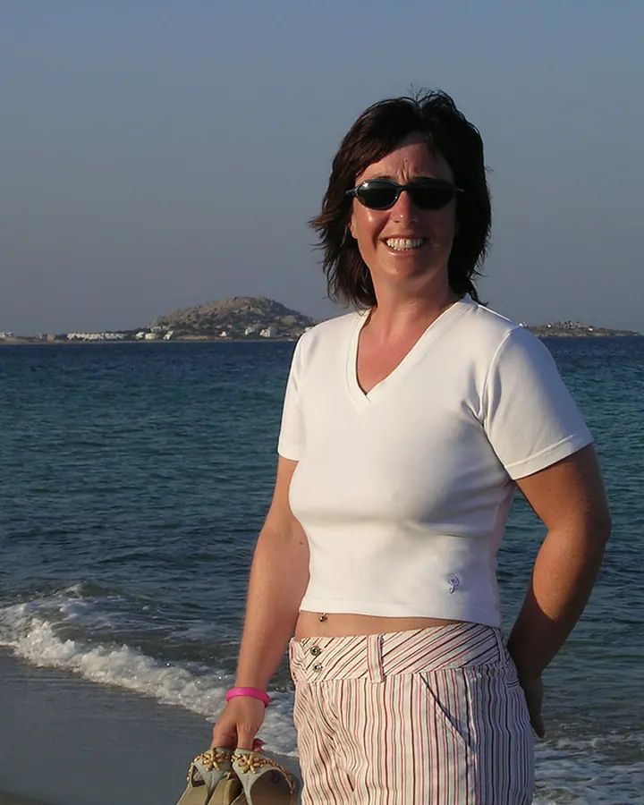 Femme aux cheveux bruns, portant un t-shirt blanc, souriant sur une plage avec la mer en arrière-plan.
