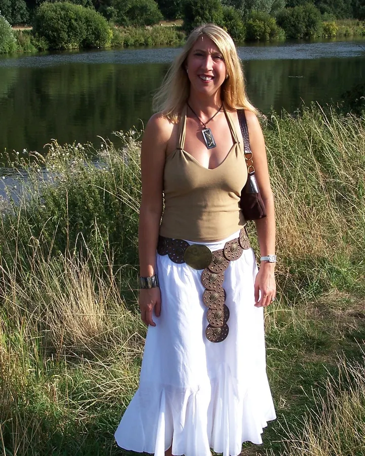 Femme blonde souriante en tenue bohème, debout près d'un lac entouré de végétation.