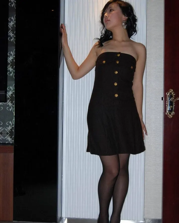 Femme asiatique aux cheveux noirs, portant une robe noire à boutons dorés, des collants noirs et des chaussures à talons, appuyée contre un mur blanc.