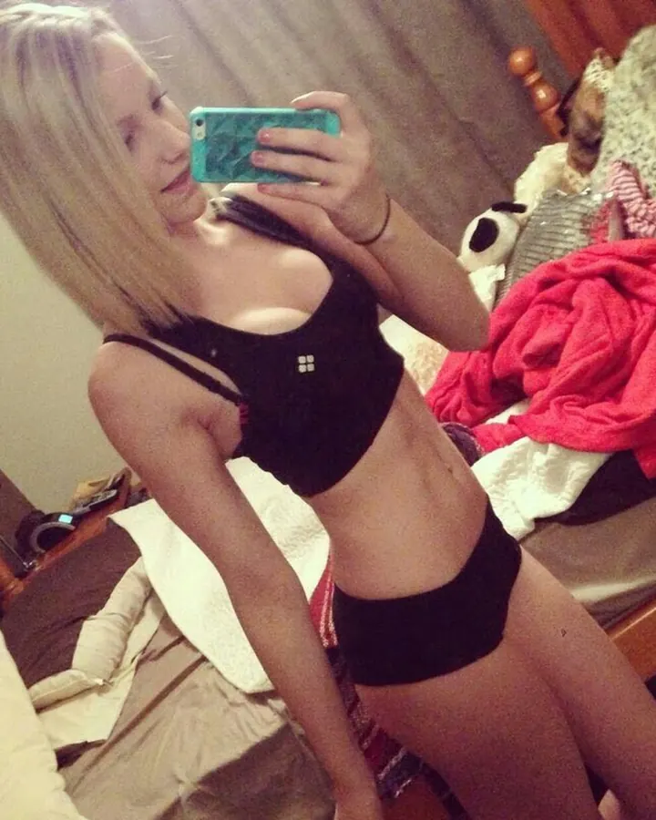 Jeune femme blonde en sous-vêtements noirs prenant un selfie devant un miroir dans une chambre désordonnée.